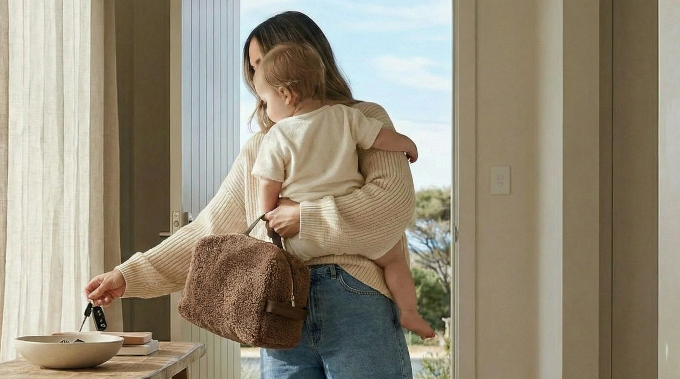 Woman holding a child and a chocolate sherpa bag at a front door ready to leave