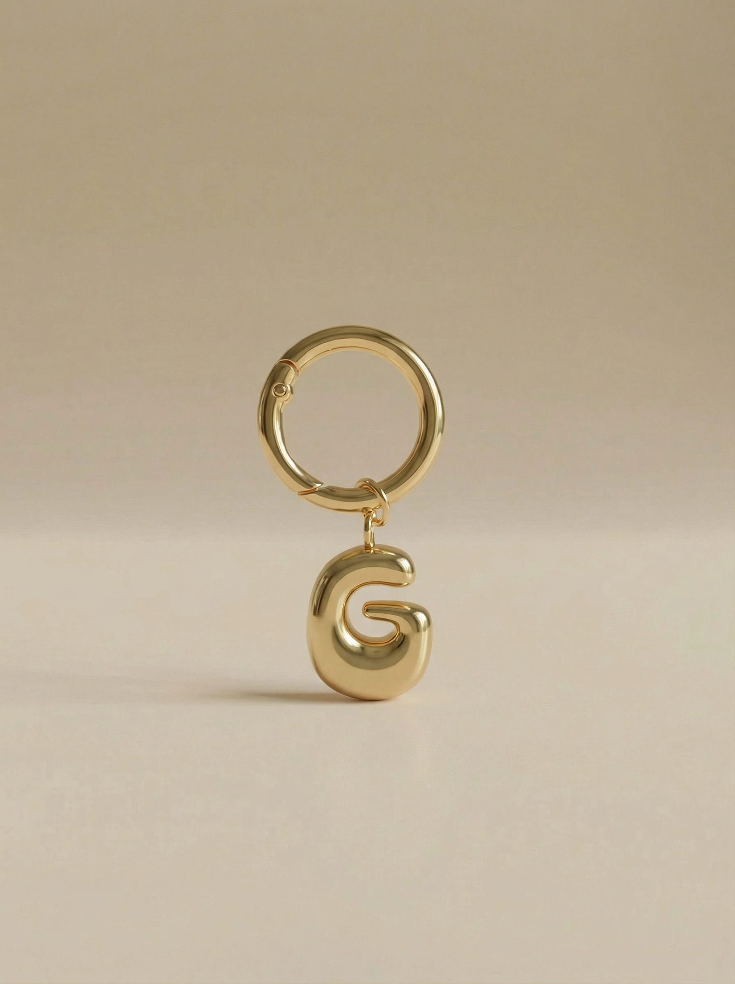 Gold charm in the letter 'G' attached to a gold keyring on beige background
