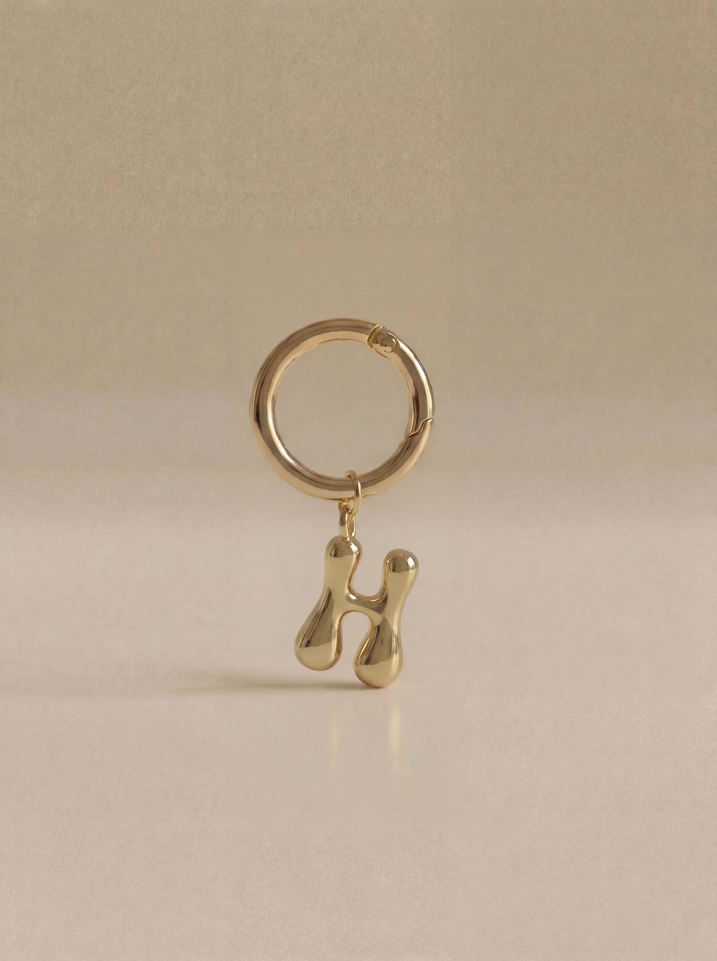 Gold charm in the letter 'H' attached to a gold keyring on beige background