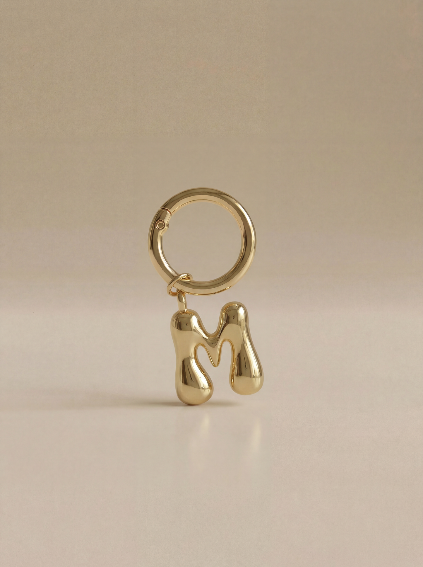 Gold charm in the letter 'M' attached to a gold keyring on beige background