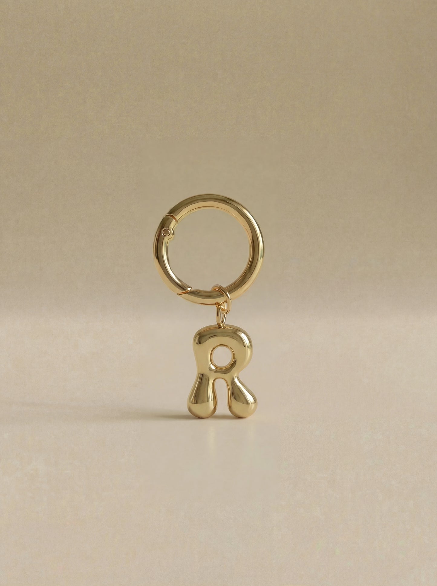 Gold charm in the letter 'R' attached to a gold keyring on beige background